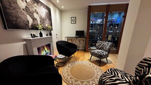 A living room with a fireplace, two black armchairs, a zebra-striped chair, a round rug, and a bookshelf with various items.