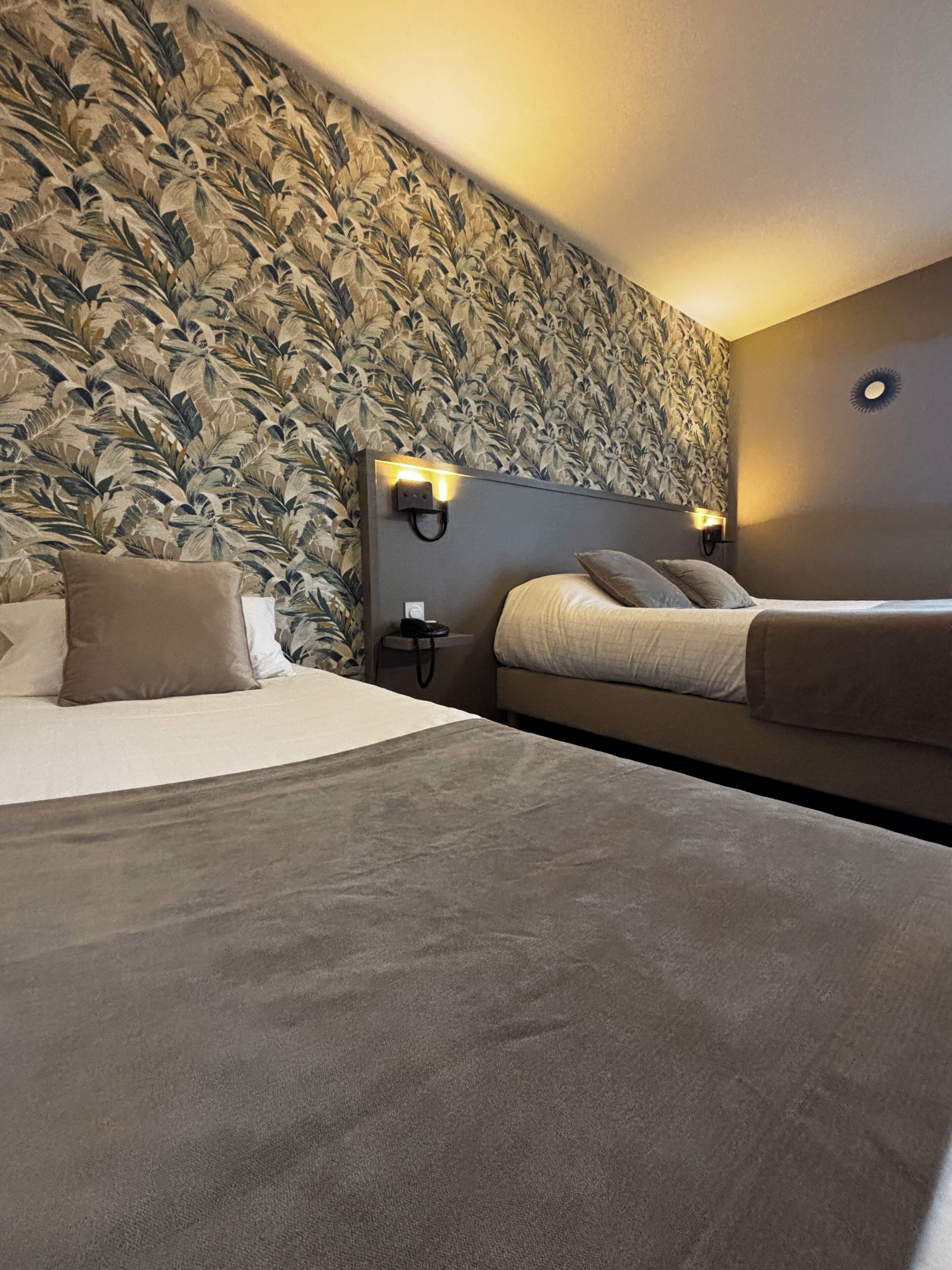A hotel room with a large bed, a smaller bed, a nightstand, and a wall with a leafy pattern.