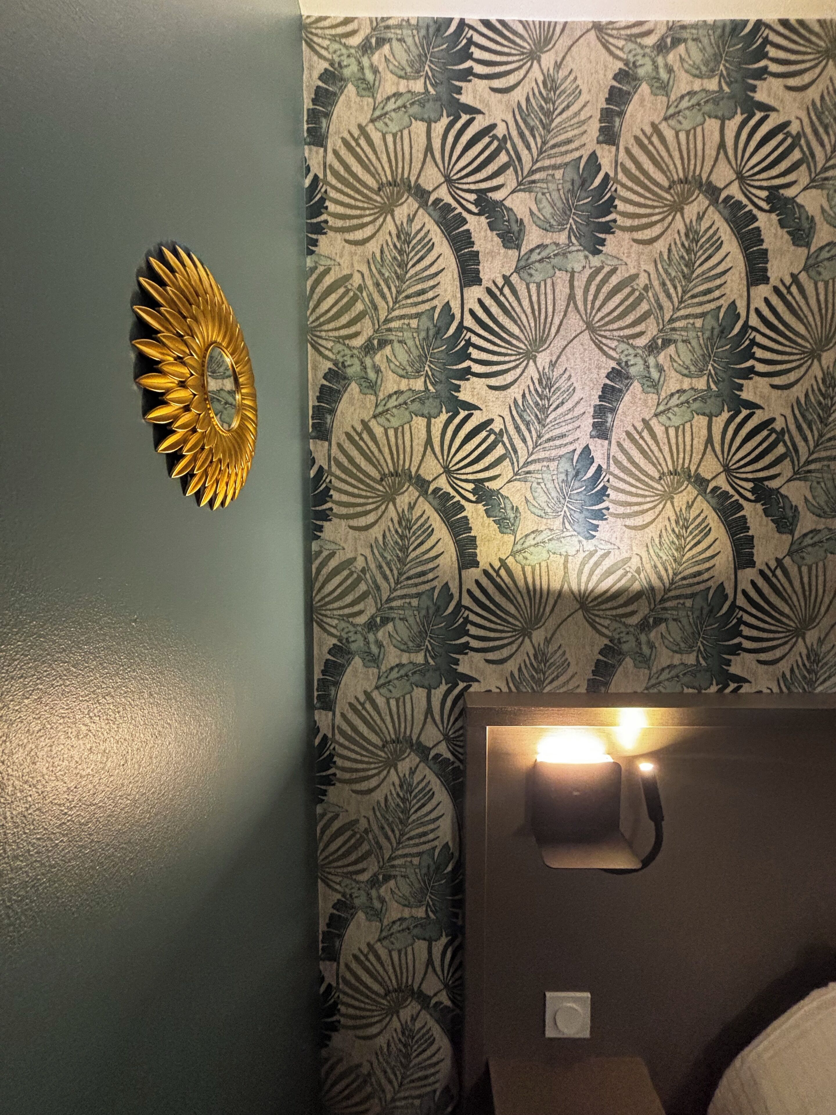 A room with a patterned wallpaper, a decorative sunburst mirror, and a bedside lamp.