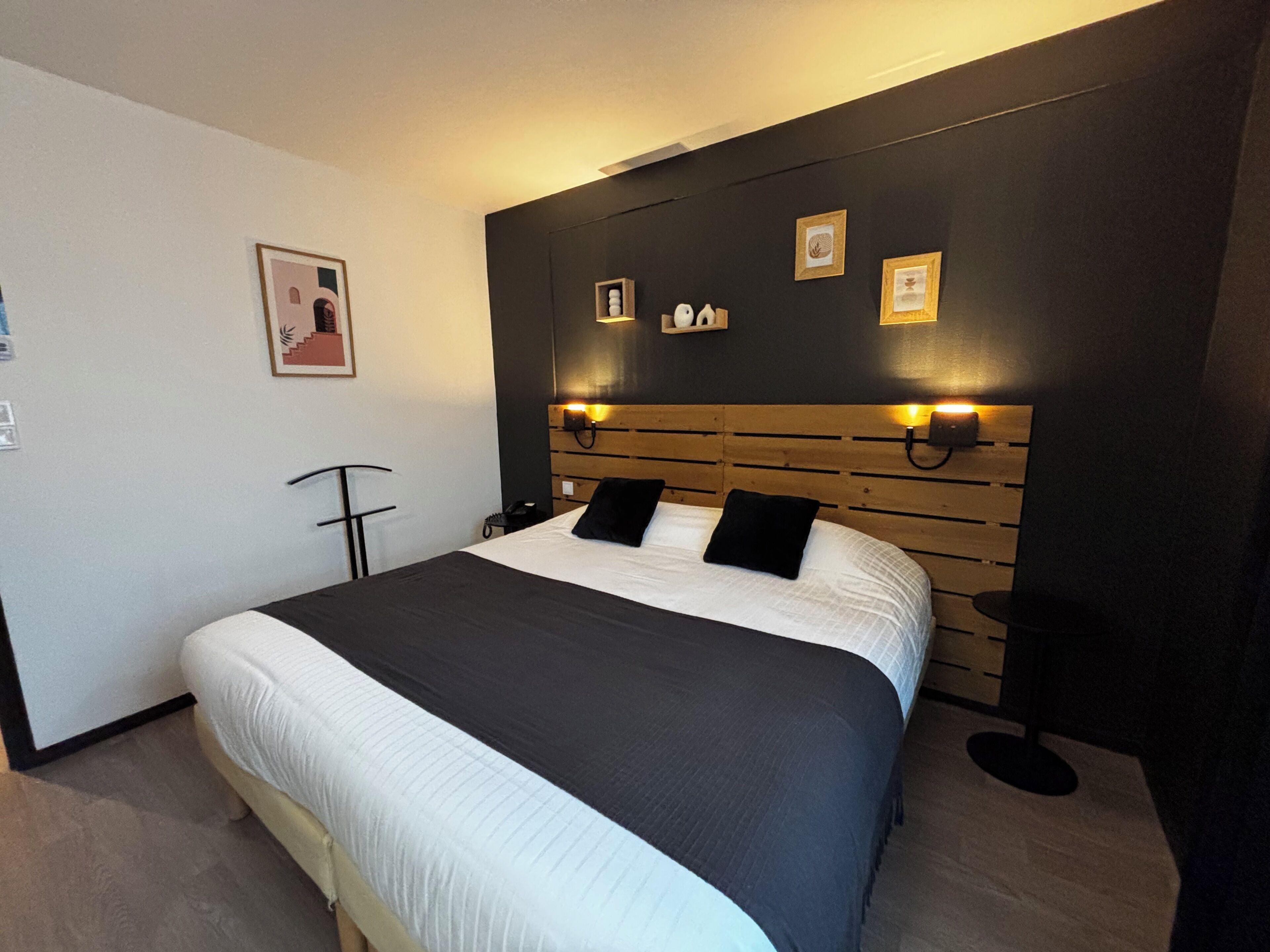 A modern hotel room with a large bed, a wooden headboard, and wall-mounted lighting.
