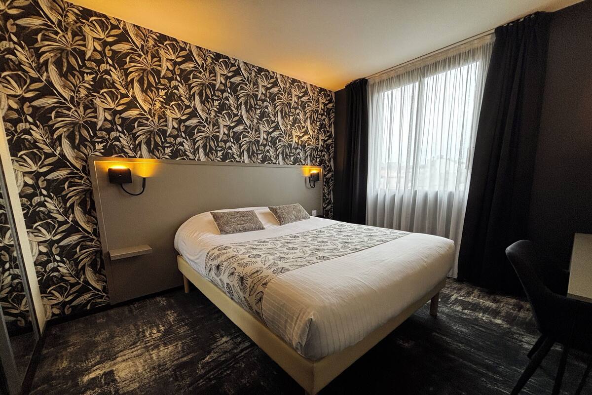 A hotel room with a large bed, a headboard with wall-mounted lights, a dark patterned wallpaper, and a view of the city through sheer curtains.