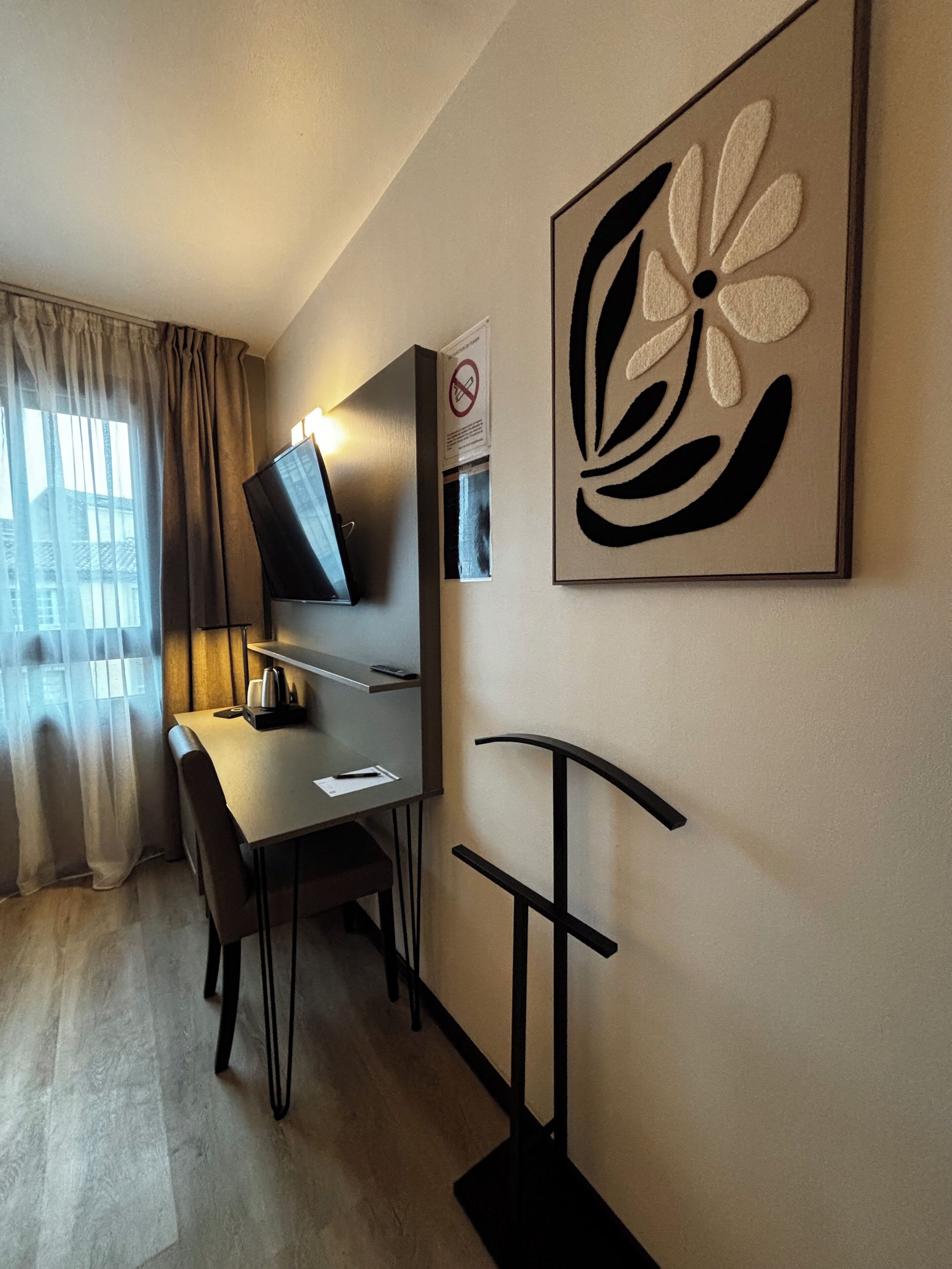 Ein Hotelzimmer mit Schreibtisch, Stuhl, Fernseher, Kunstwerken und einem Fenster mit Vorhängen.