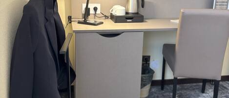 A compact hotel room with a kitchenette, a flat-screen TV mounted on the wall, a desk with a lamp, a chair, and a coat rack.