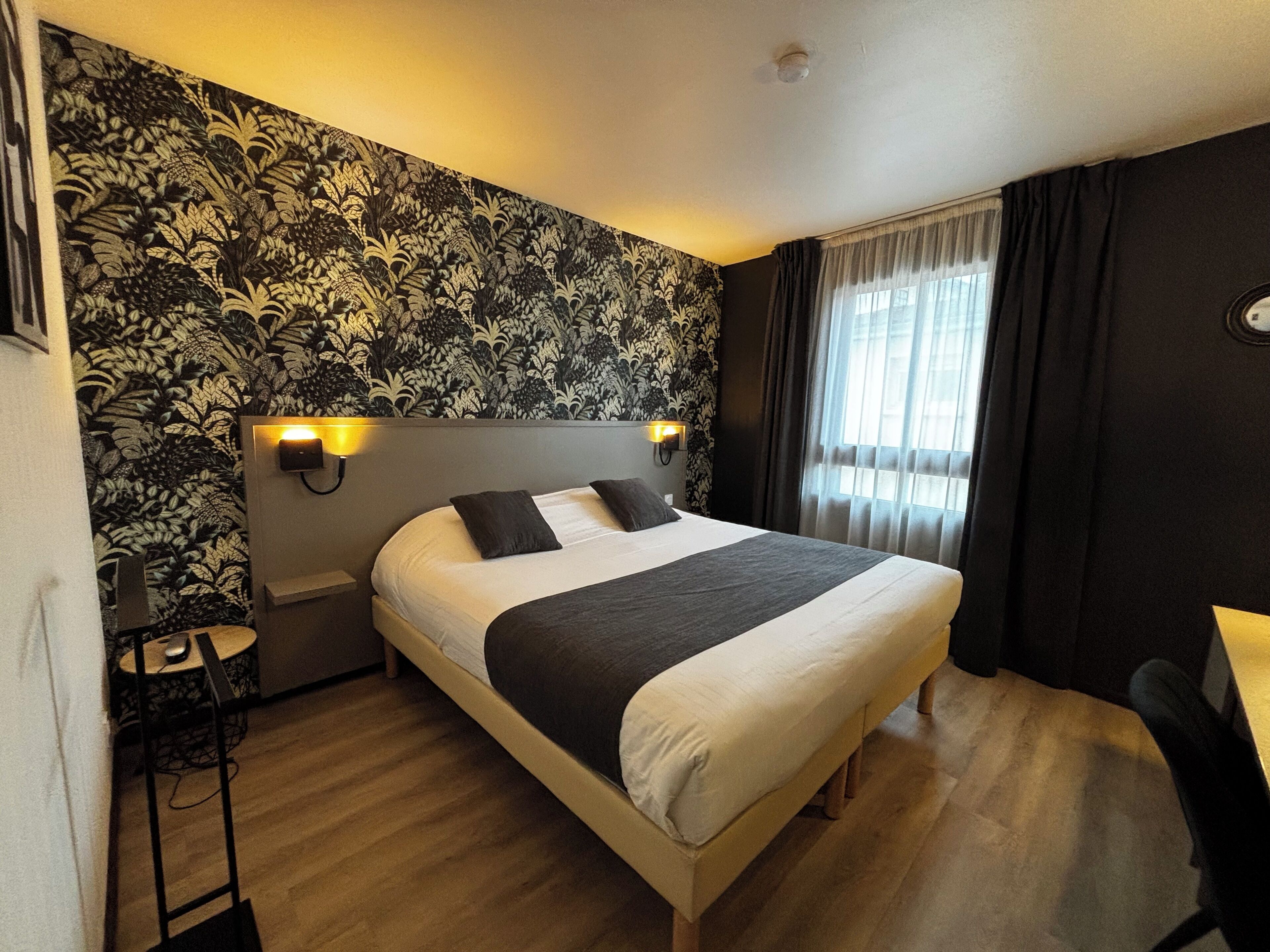 A hotel room with a large bed, two bedside lamps, a small desk with a chair, and a window with curtains.