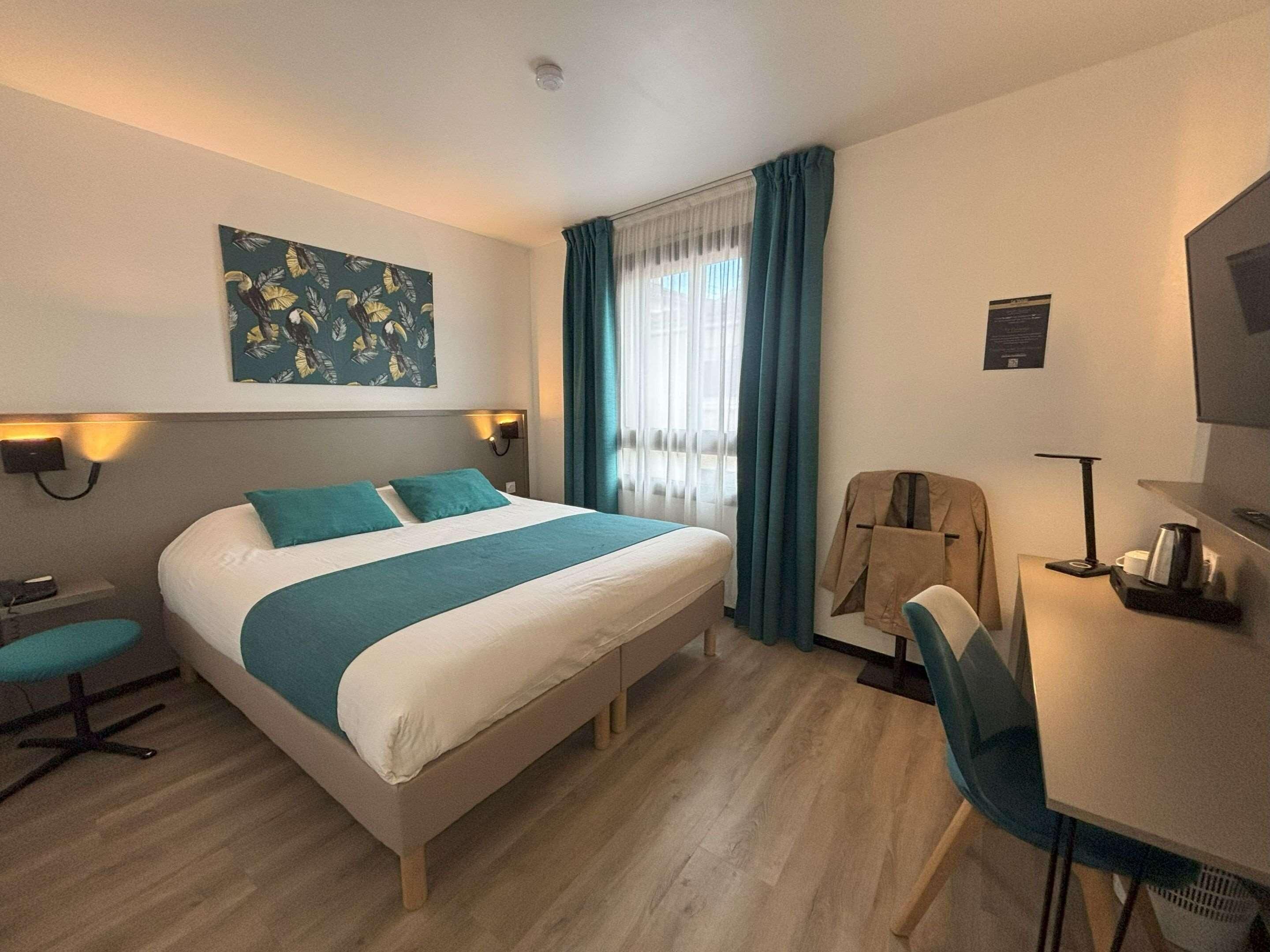 A hotel room with a bed, a desk, a chair, a coat rack, and a painting on the wall.