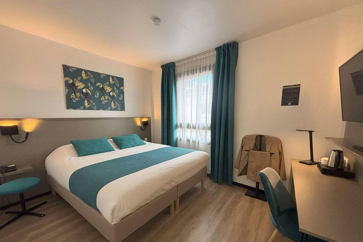 A hotel room with a bed, a desk, a chair, a coat rack, and a painting on the wall.