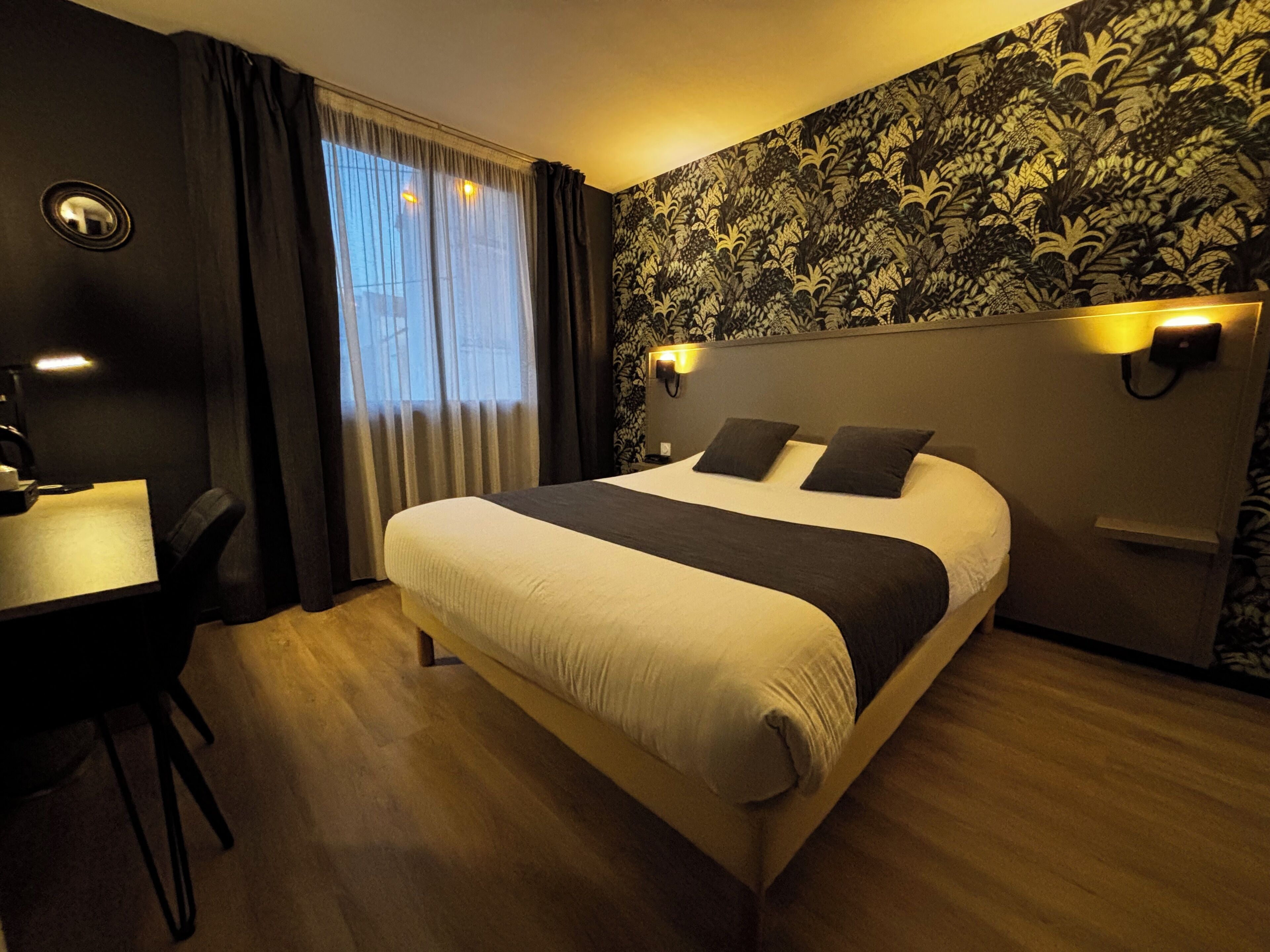 Habitación de hotel con cama, escritorio y silla. Se ve una ventana con cortinas, una pared con estampado floral y una lámpara de pared.