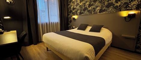 Habitación de hotel con cama, escritorio y silla. Se ve una ventana con cortinas, una pared con estampado floral y una lámpara de pared.