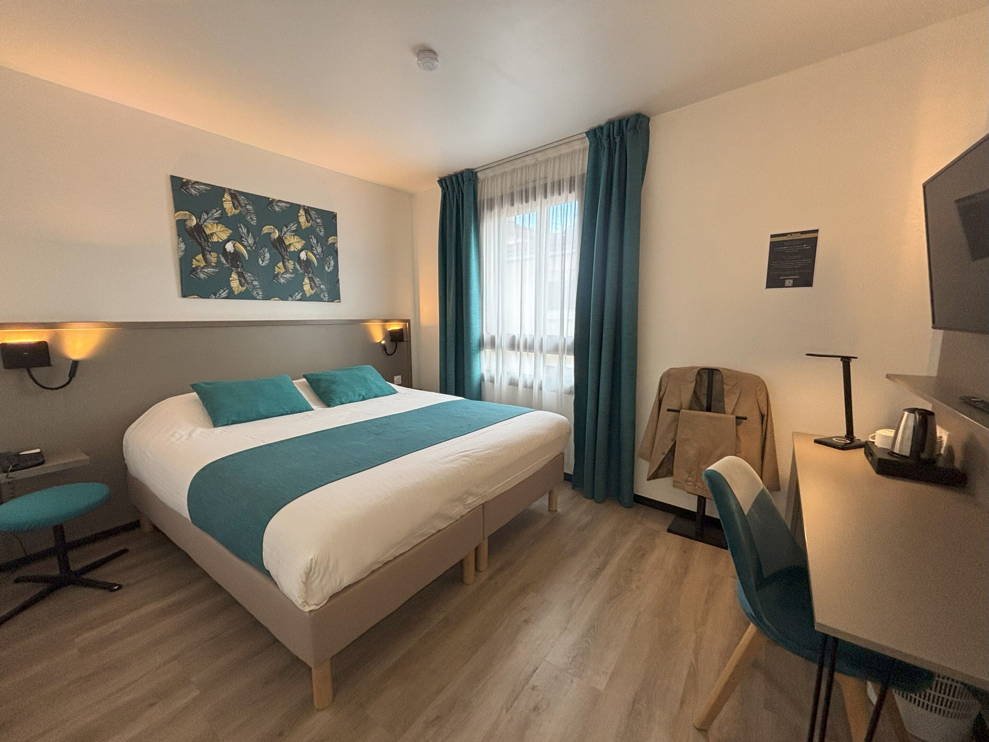 Habitación de hotel con cama, escritorio, silla, perchero y un cuadro en la pared.