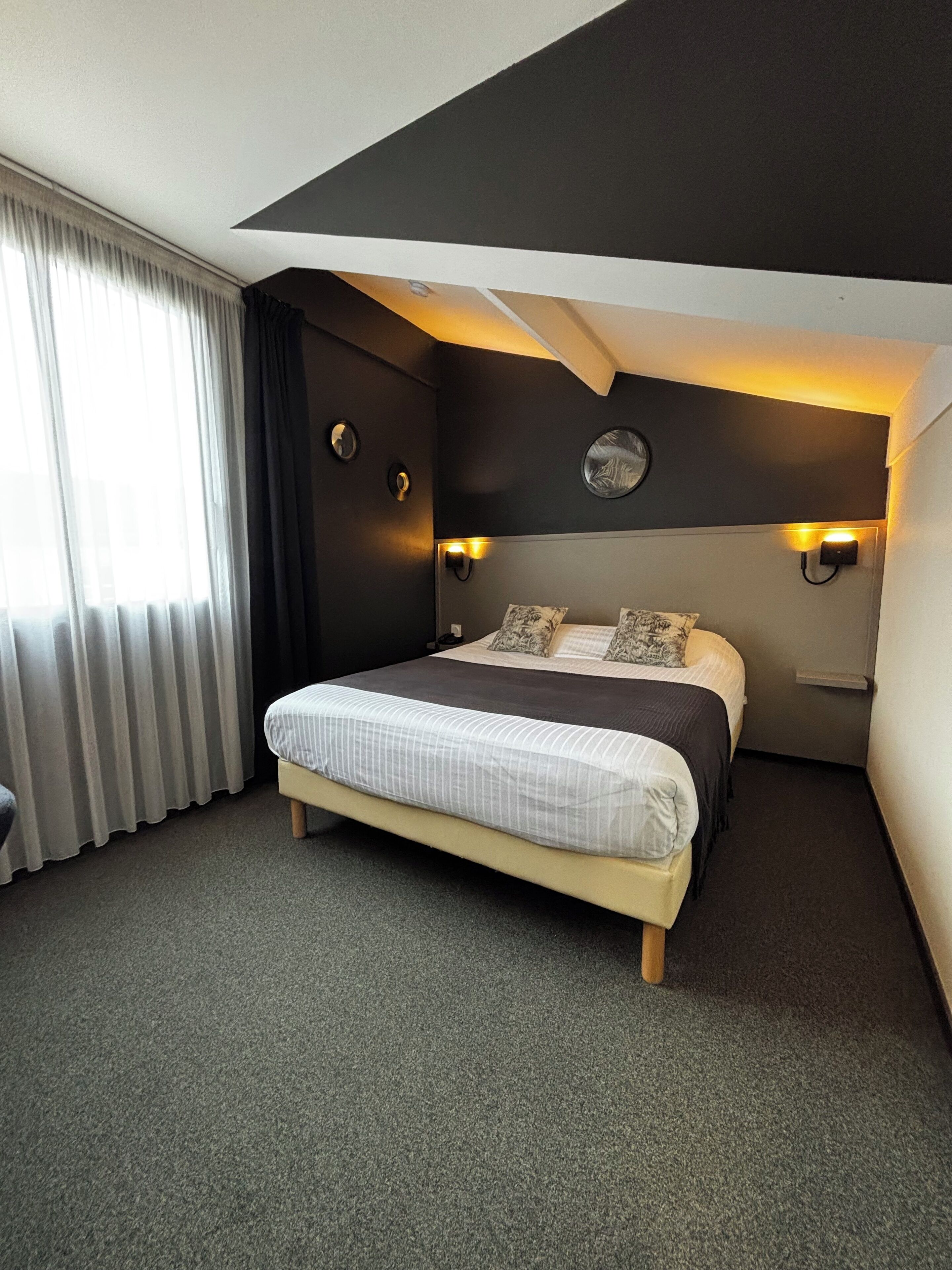 Quarto de hotel moderno com cama grande, paredes escuras e duas luminárias fixadas na parede.