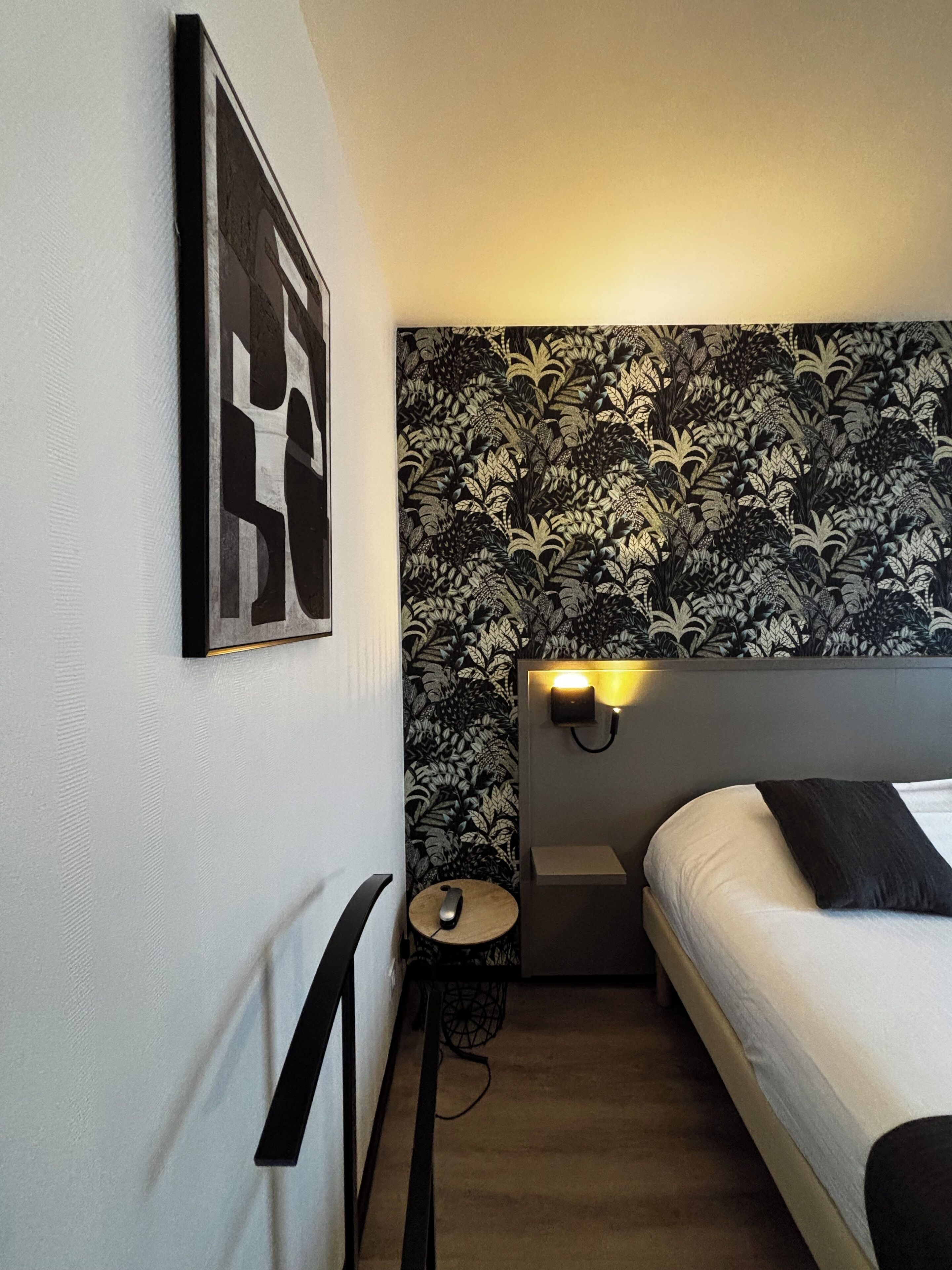 A bedroom with a bed, a headboard with a light, a small table with a remote, and a wall with a floral wallpaper.