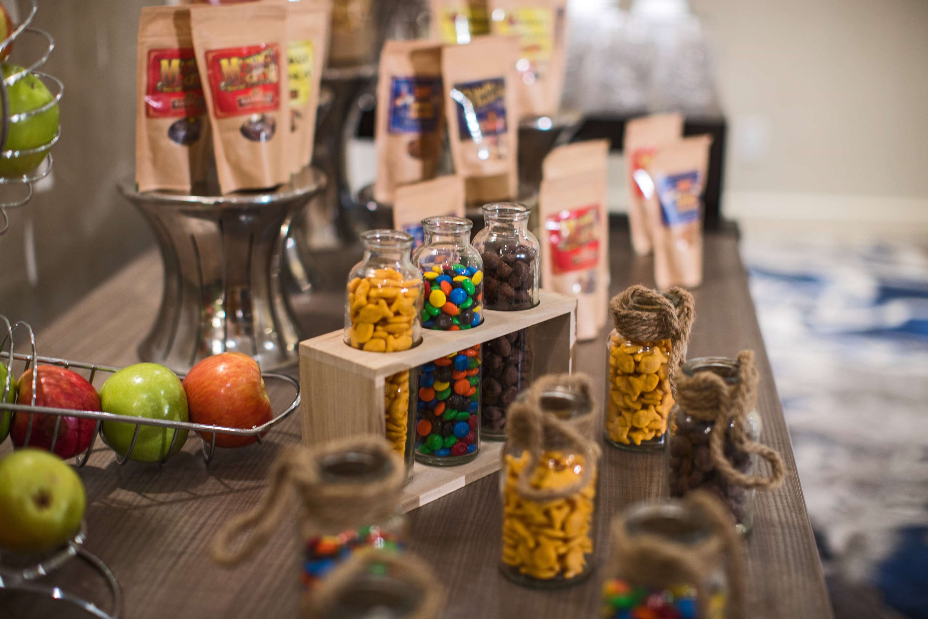 Ein Tisch mit verschiedenen Snacks und Süßigkeiten in Gläsern, ein Drahtobstkorb mit Äpfeln und Papierbeutel im Hintergrund.
