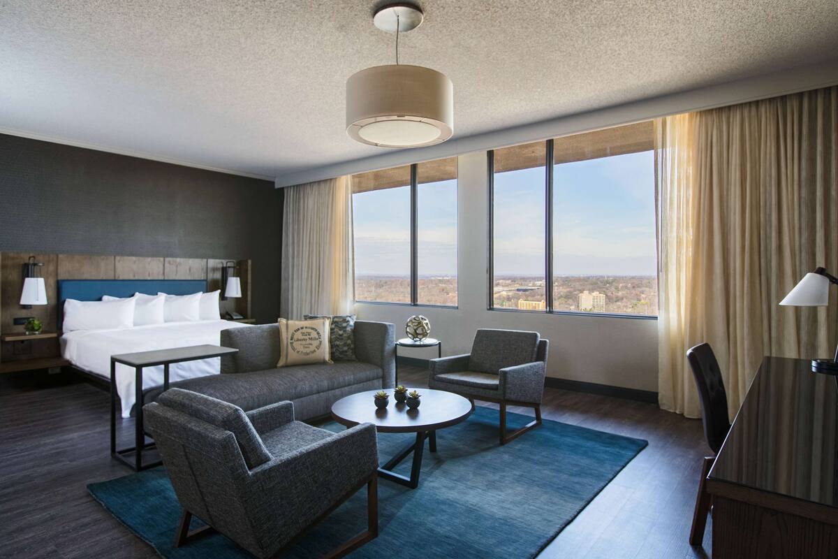 A hotel room with a bed, sofa, chairs, a coffee table, and a view of the city.