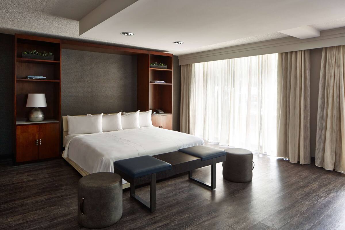 A modern hotel room with a large bed, built-in shelving, a bench, and a large window with curtains.