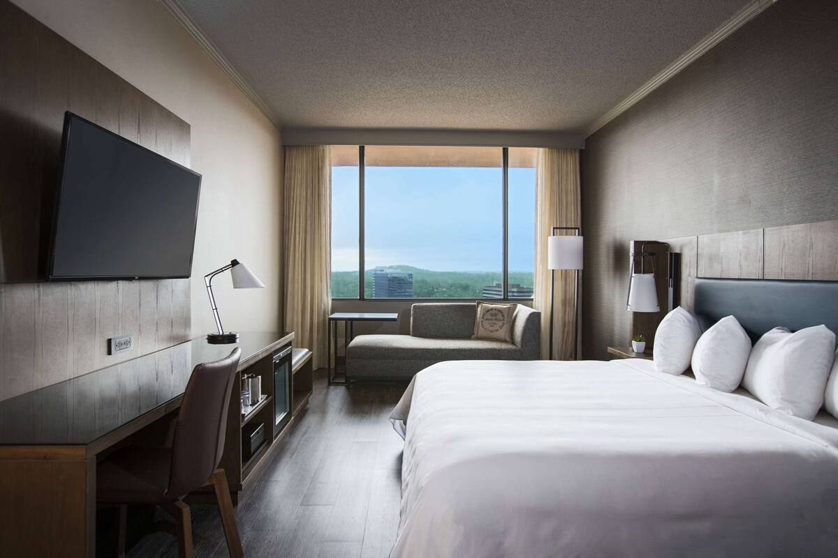 A hotel room with a large bed, a desk with a chair, a television, a sofa, and a window with a city view.