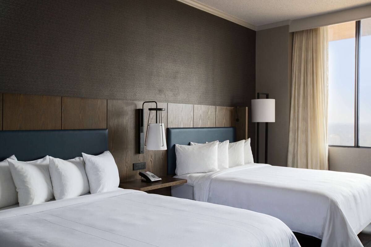 A hotel room with two beds, a nightstand with a lamp, a telephone, and a window with curtains.