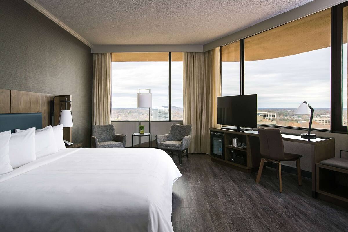 A hotel room with a large bed, a desk with a TV, two chairs, a small table with a lamp, and a view of the city through large windows.