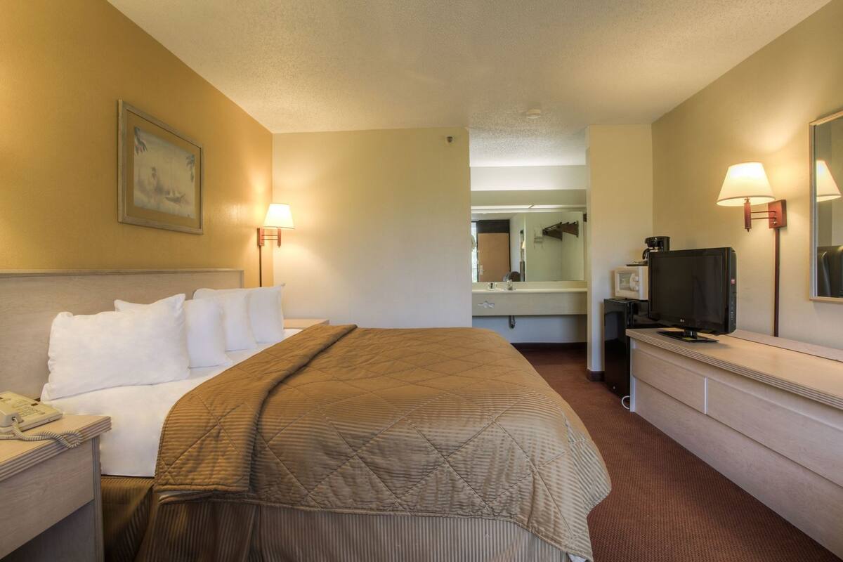 Una habitación de hotel con cama, mesita de noche, lámpara, televisión y espejo.