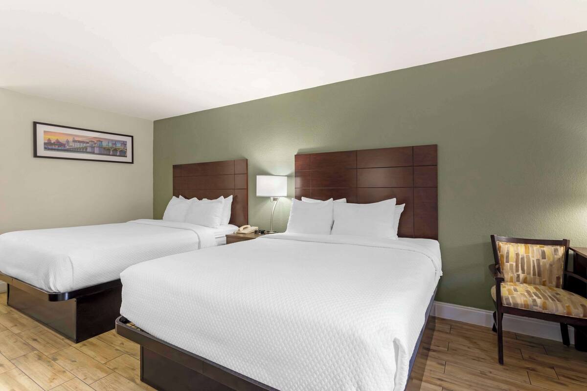 Una habitación de hotel con dos camas, una mesita de noche con lámpara, una silla y un cuadro en la pared.