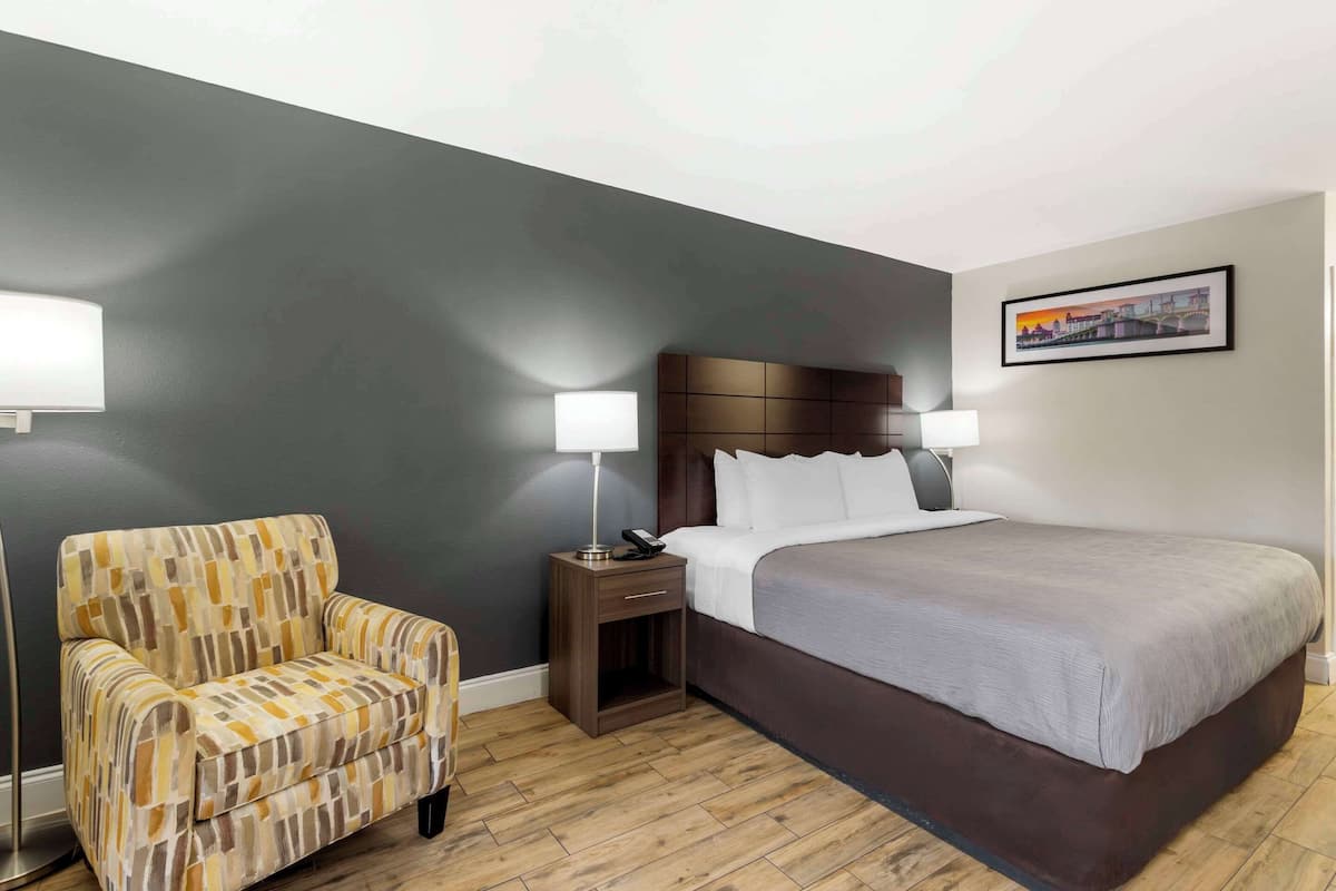 Una habitación de hotel con una cama, una silla, una mesita de noche con una lámpara y un cuadro en la pared.
