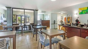 Un comedor con mesas y sillas, una zona de cocina con electrodom ésticos y vistas a través de un ventanal.