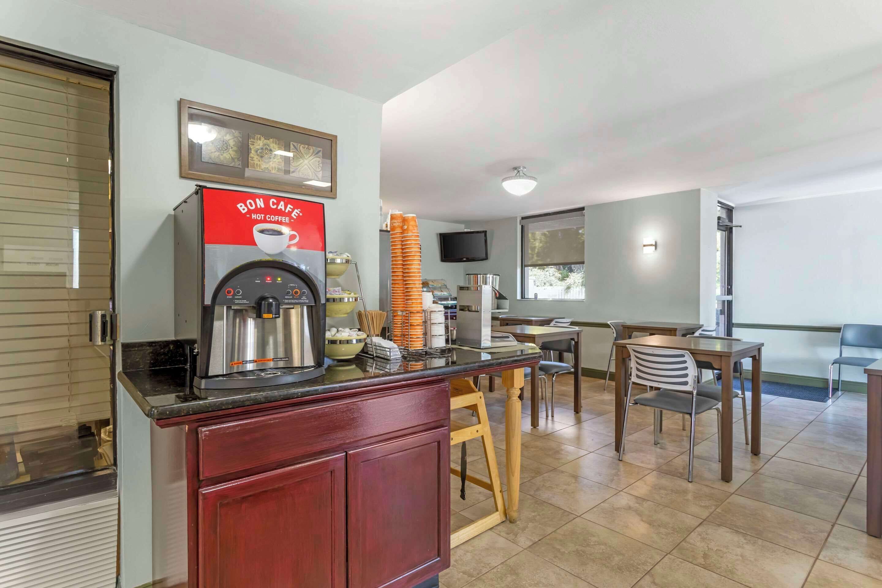 Una kitchenette con una cafetera, una pila de vasos naranjas y un comedor con sillas y una mesa.