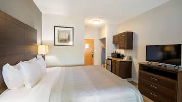 A hotel room with a large bed, a TV, a desk with a lamp, and a kitchen area.