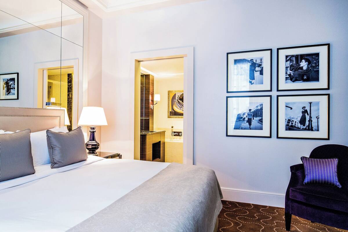 Hotellihuone, jossa on suuri sänky, tuoli, yöpöytä lampun kanssa ja seinällä kehystettyjä kuvia.