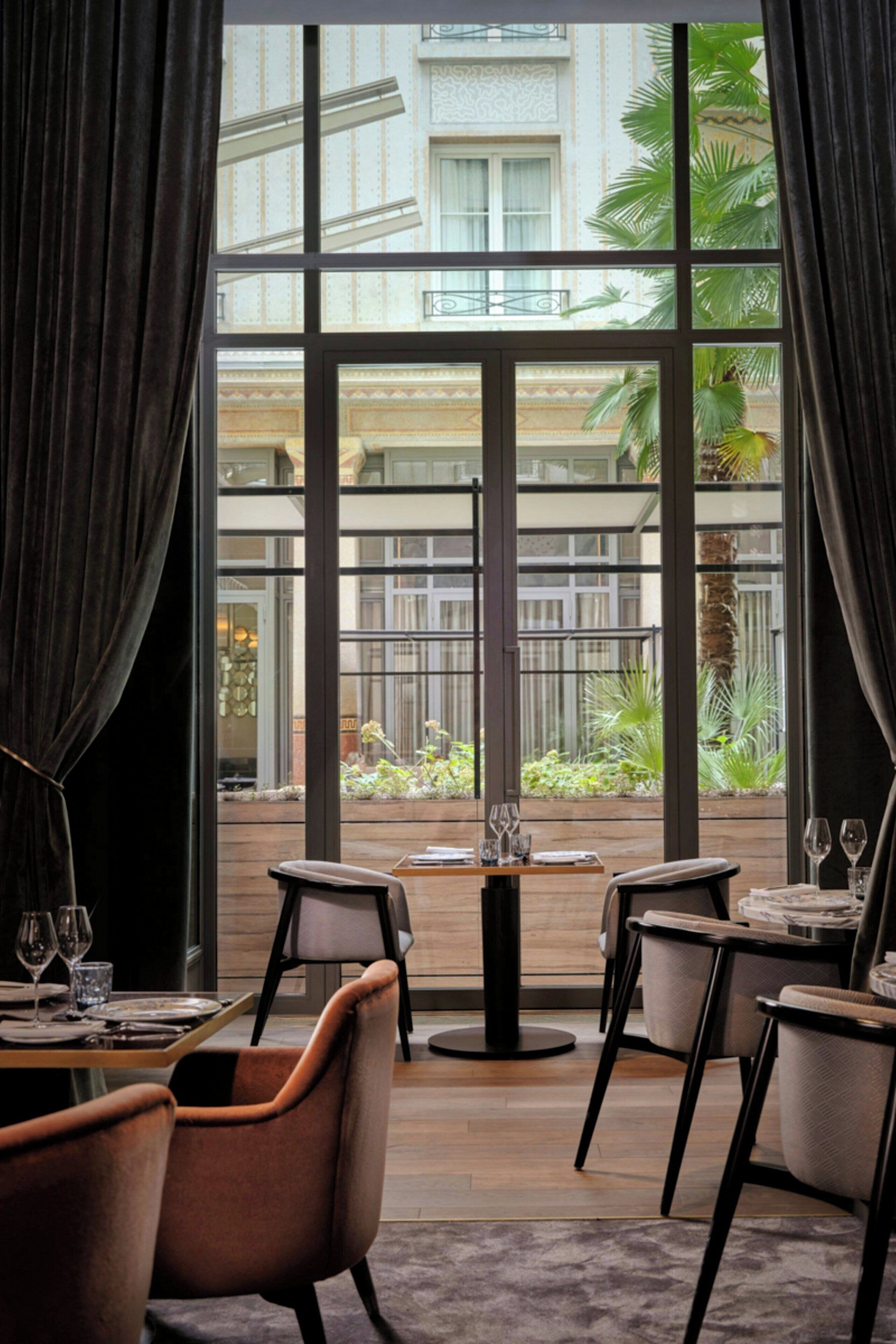 A dining area with a table set for two, large windows, and a view of a building with balconies.