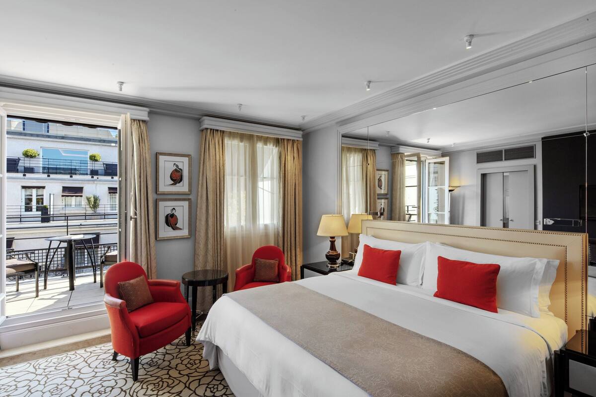 Hotellihuone, jossa on suuri sänky, punainen nojatuoli, pieni pöytä ja parveke, jolta on näkymä kaupunkiin.