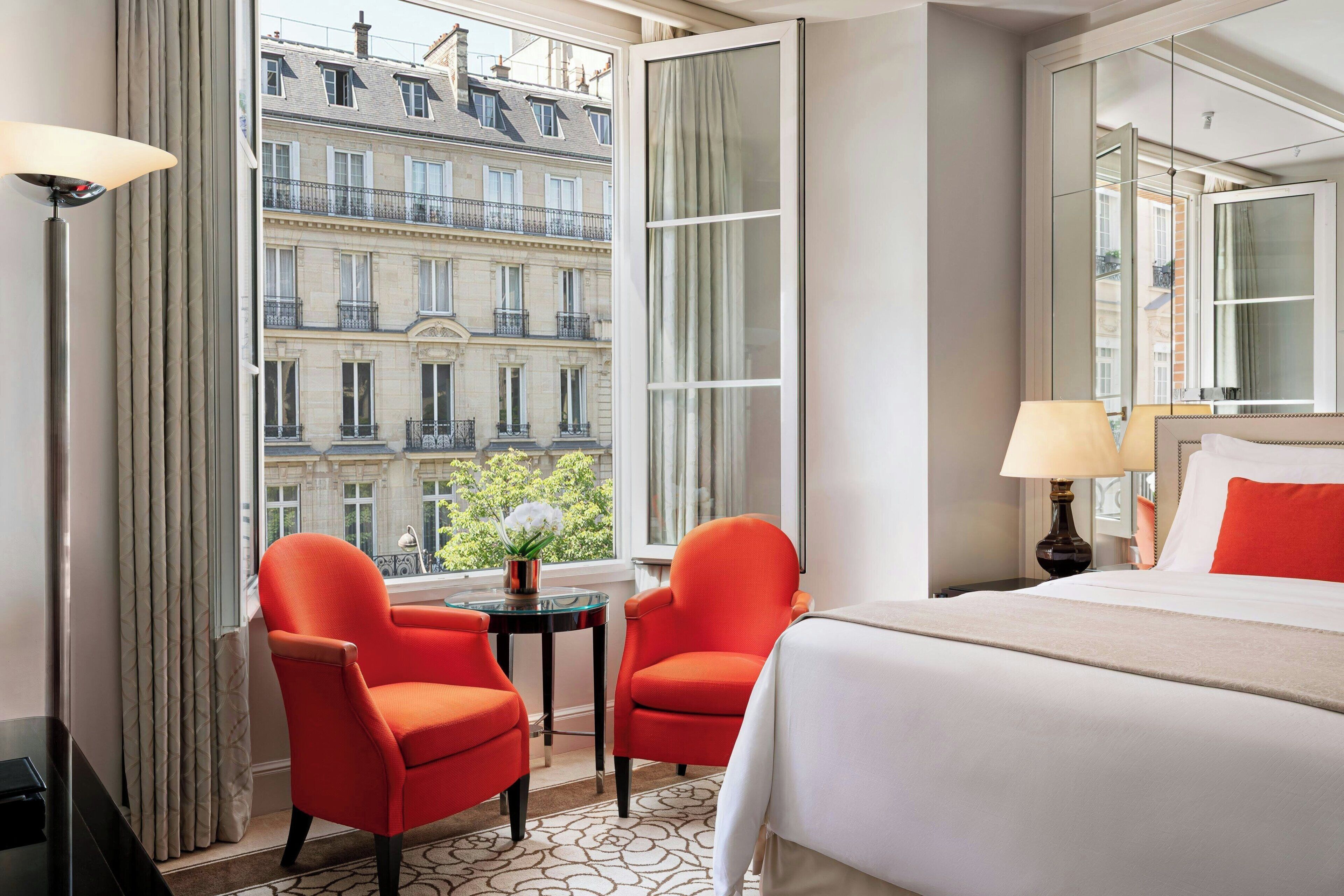 A hotel room with a large bed, two red chairs, a small table, and a view of a building with balconies.