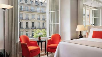 A hotel room with a large bed, two red chairs, a small table, and a view of a building with balconies.