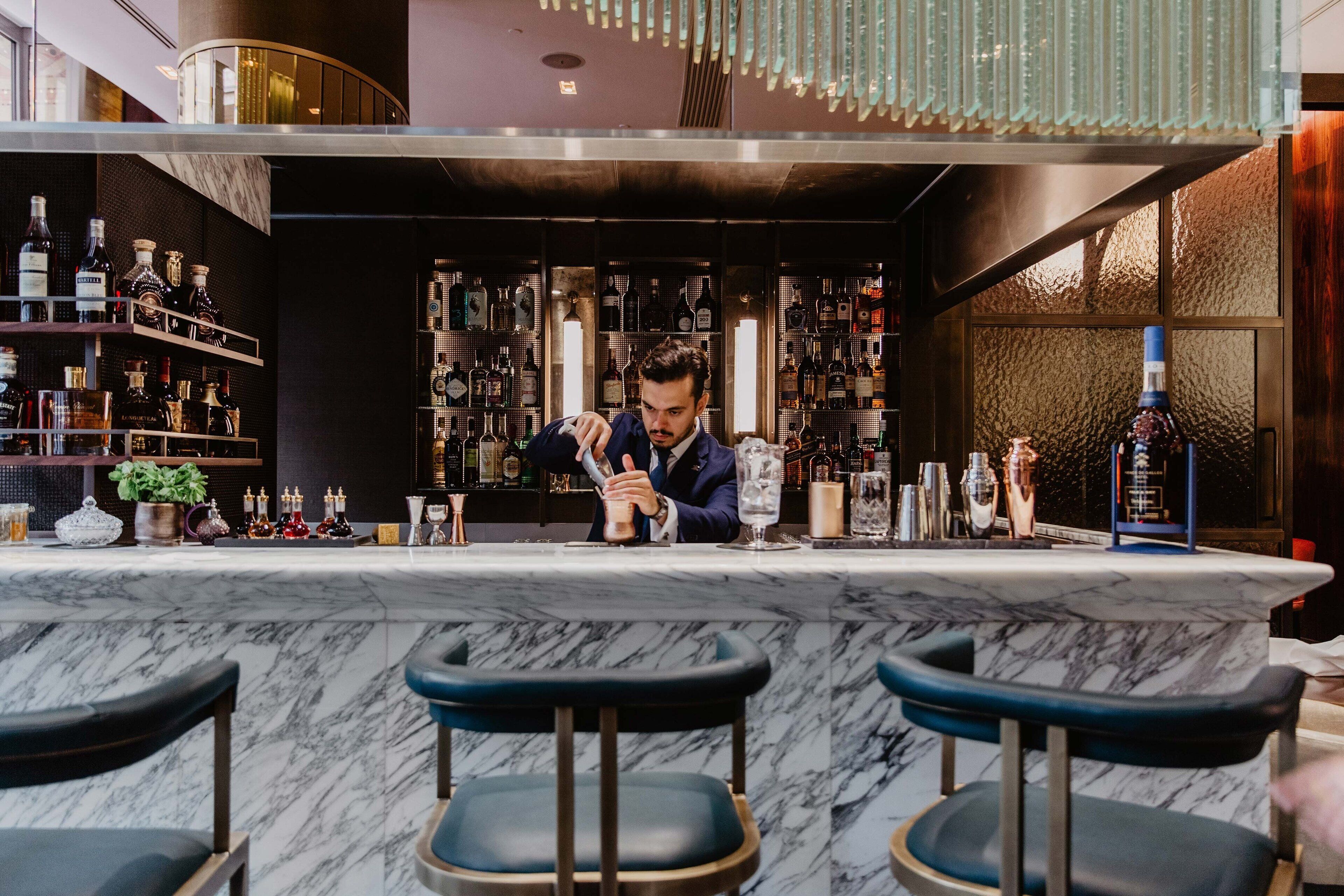 Una zona de bar con mostrador de mármol, varias estanterías llenas de botellas de licores y tres taburetes.