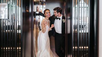 Un couple, vêtu de manière élégante, se trouve dans un ascenseur dont l’intérieur présente un design moderne, avec notamment des stores verticaux et un sol réfléchissant.