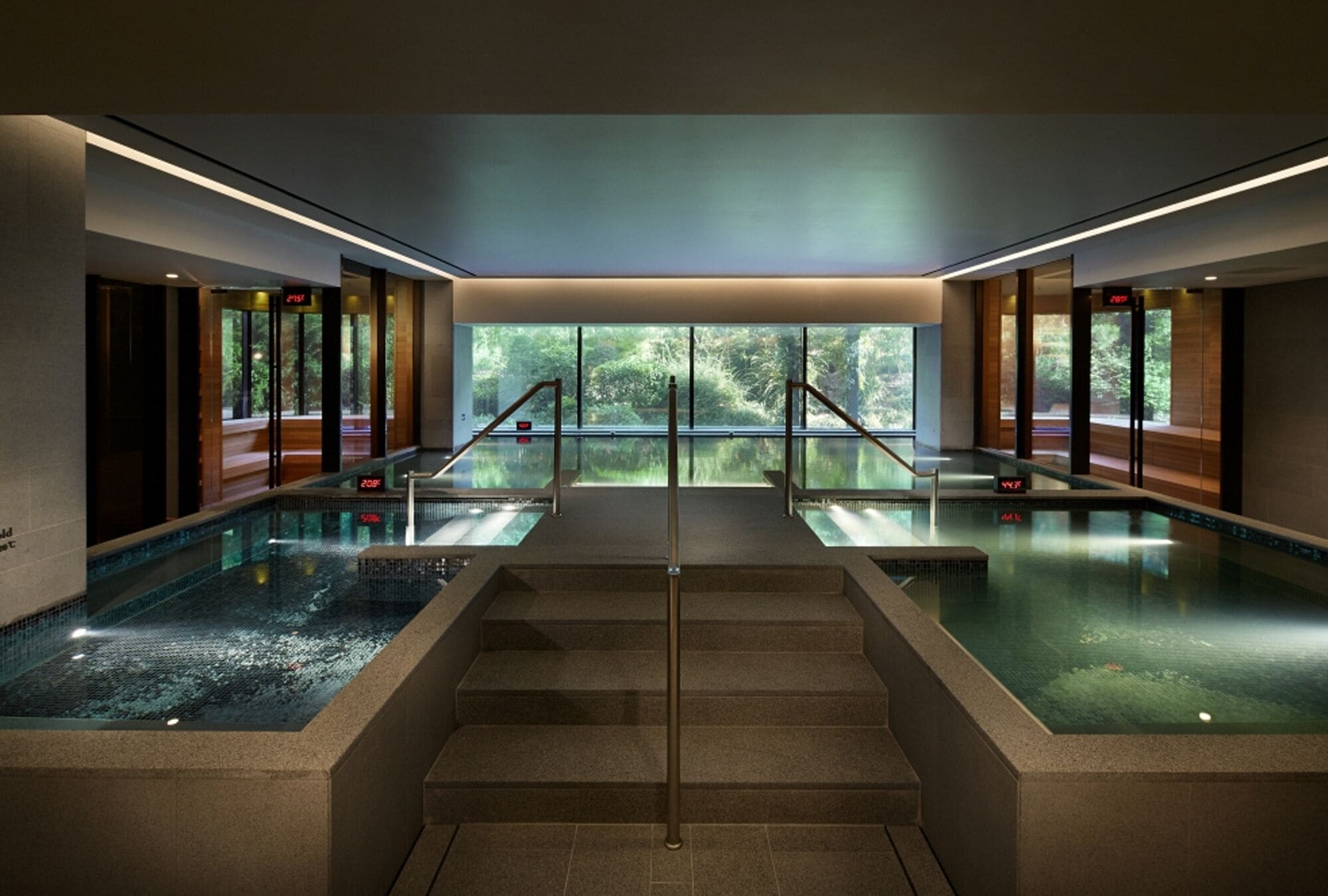 A modern indoor pool area with glass walls, a central staircase, and a view of the outdoors.