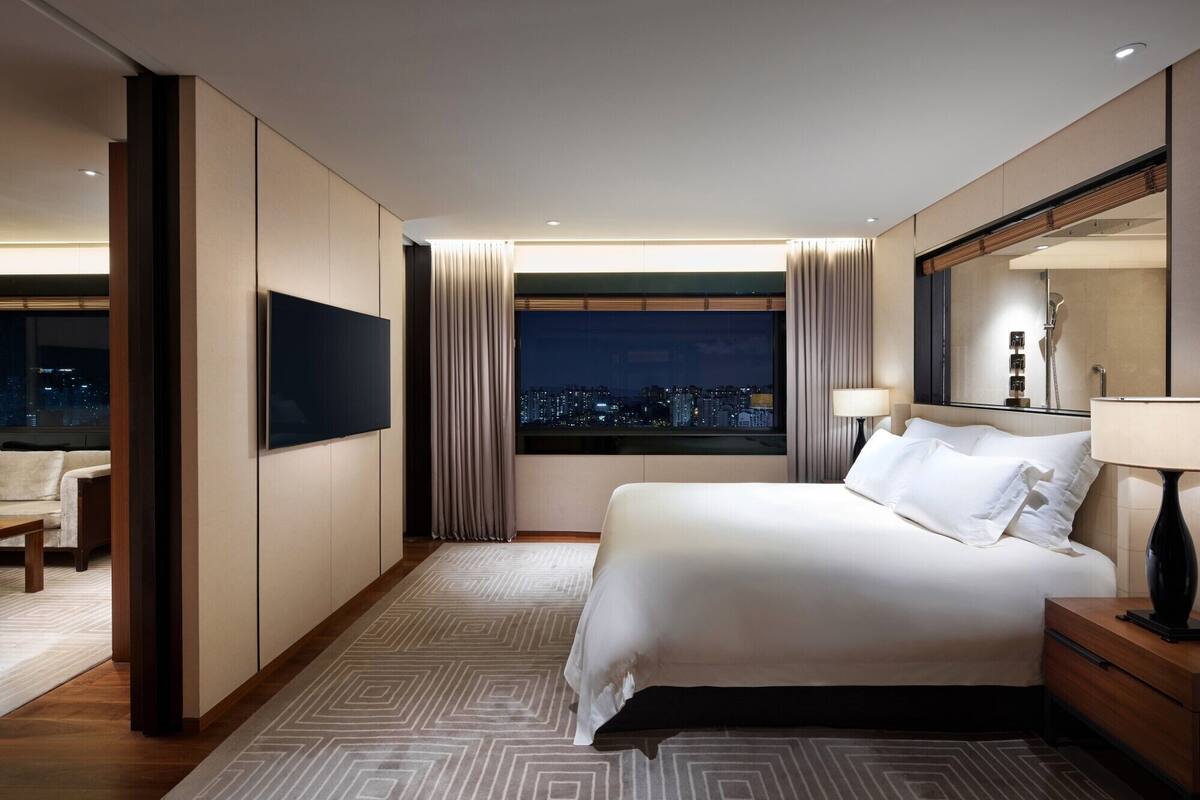 Quarto de hotel com cama, televisão, mesa de cabeceira, abajur e vista da cidade à noite.