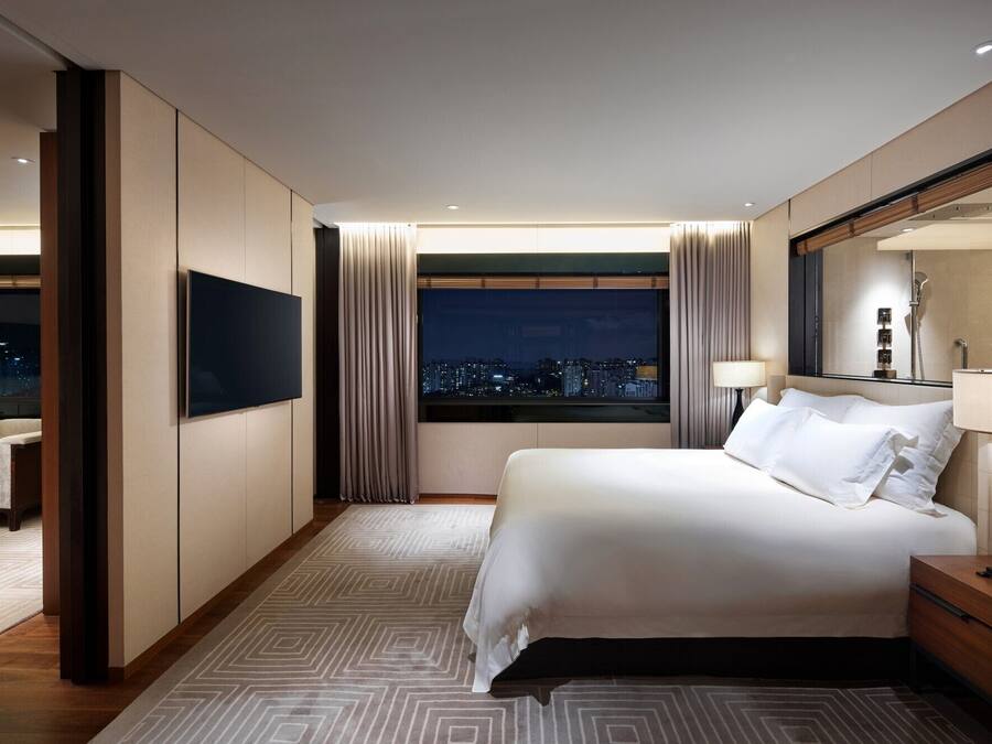 Ein Hotelzimmer mit Bett, Fernseher, Nachttisch, Lampe und Blick auf die beleuchtete Stadt.