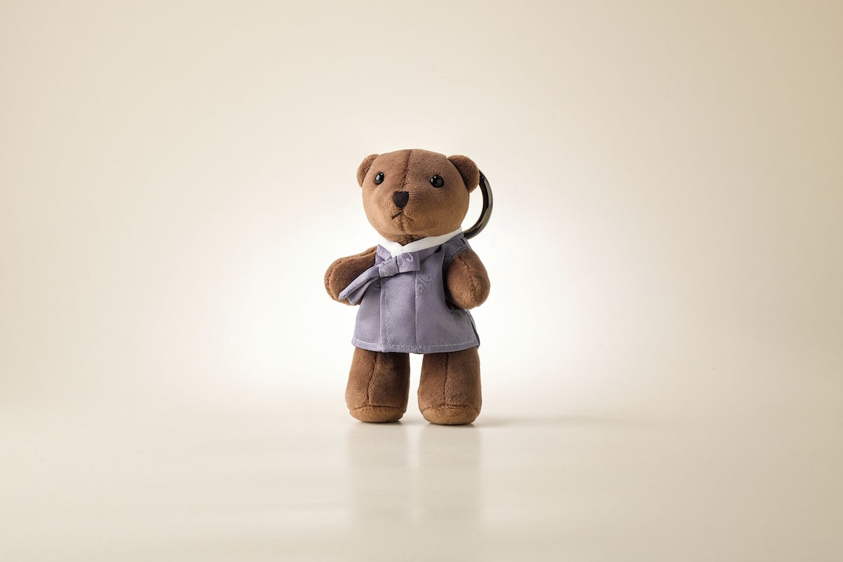 Um pequeno urso de pelúcia marrom com gravata borboleta e fita preta, em pé diante de um fundo liso.