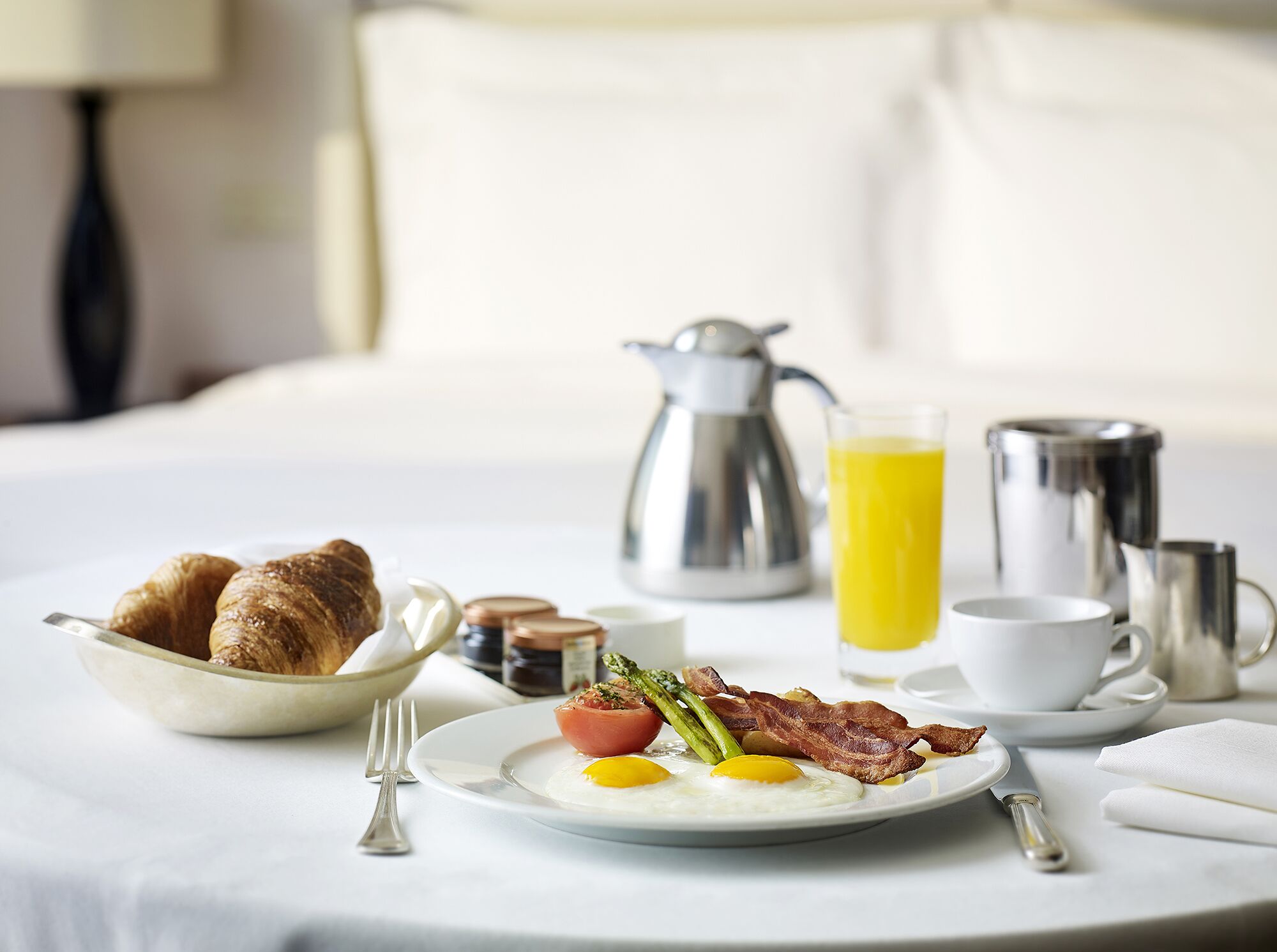 A breakfast setup on a bed with croissants, eggs, bacon, asparagus, and a glass of orange juice.