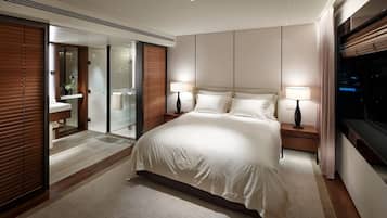 A modern hotel room with a large bed, two bedside tables with lamps, a glass shower enclosure, and a view of the city at night.