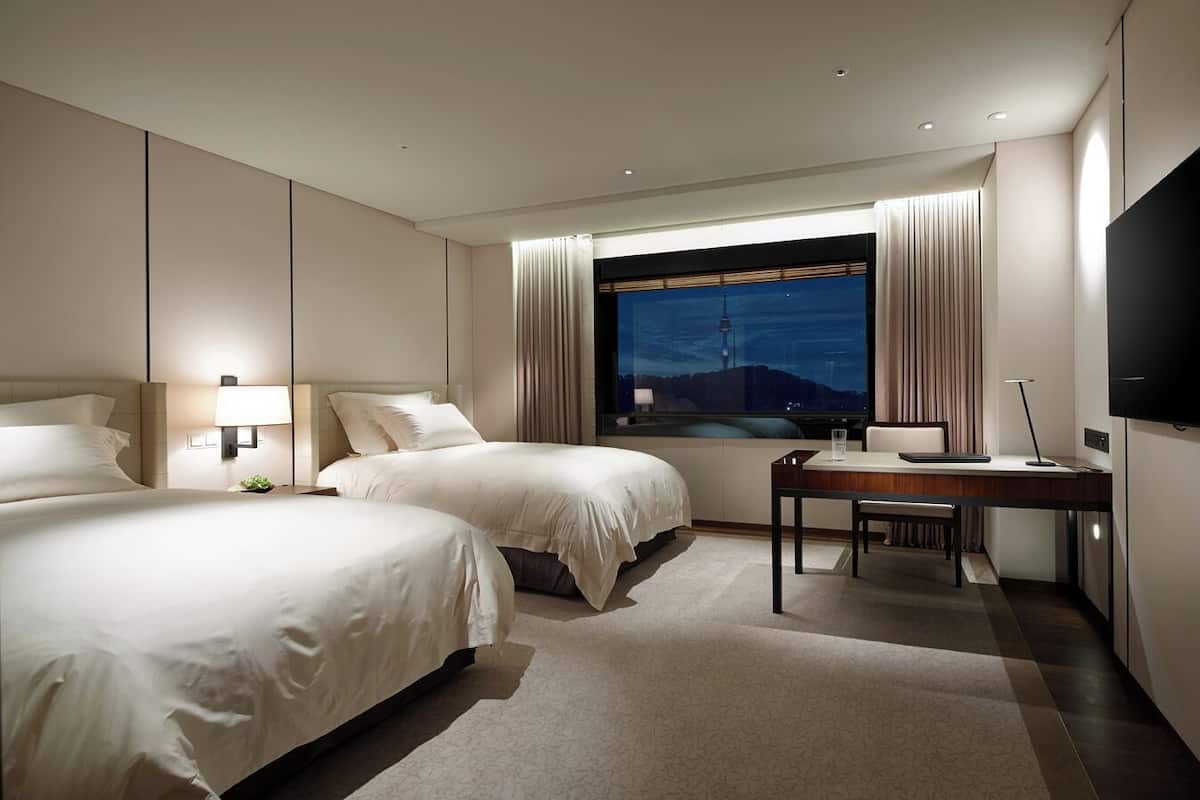 Quarto de hotel com duas camas, uma escrivaninha, televisão e janela com vista para a cidade.