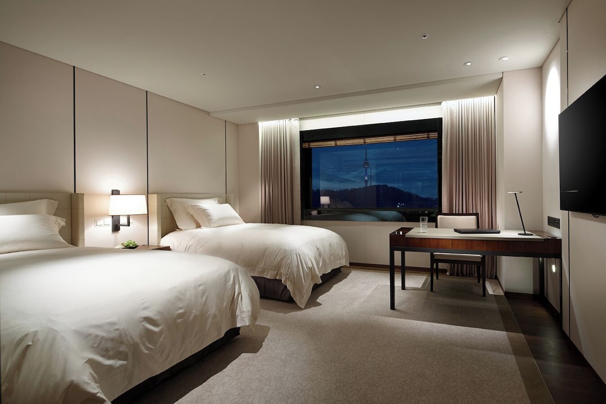 A hotel room with two beds, a desk, a TV, and a window with a cityscape view.