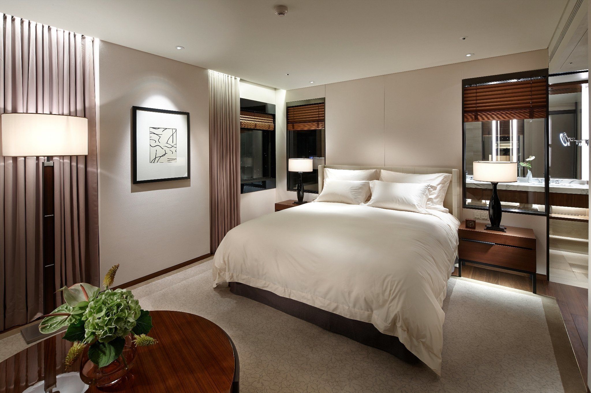 Una habitación de hotel moderna con una cama grande, lámparas de noche, un espejo y una pequeña mesa con un florero.