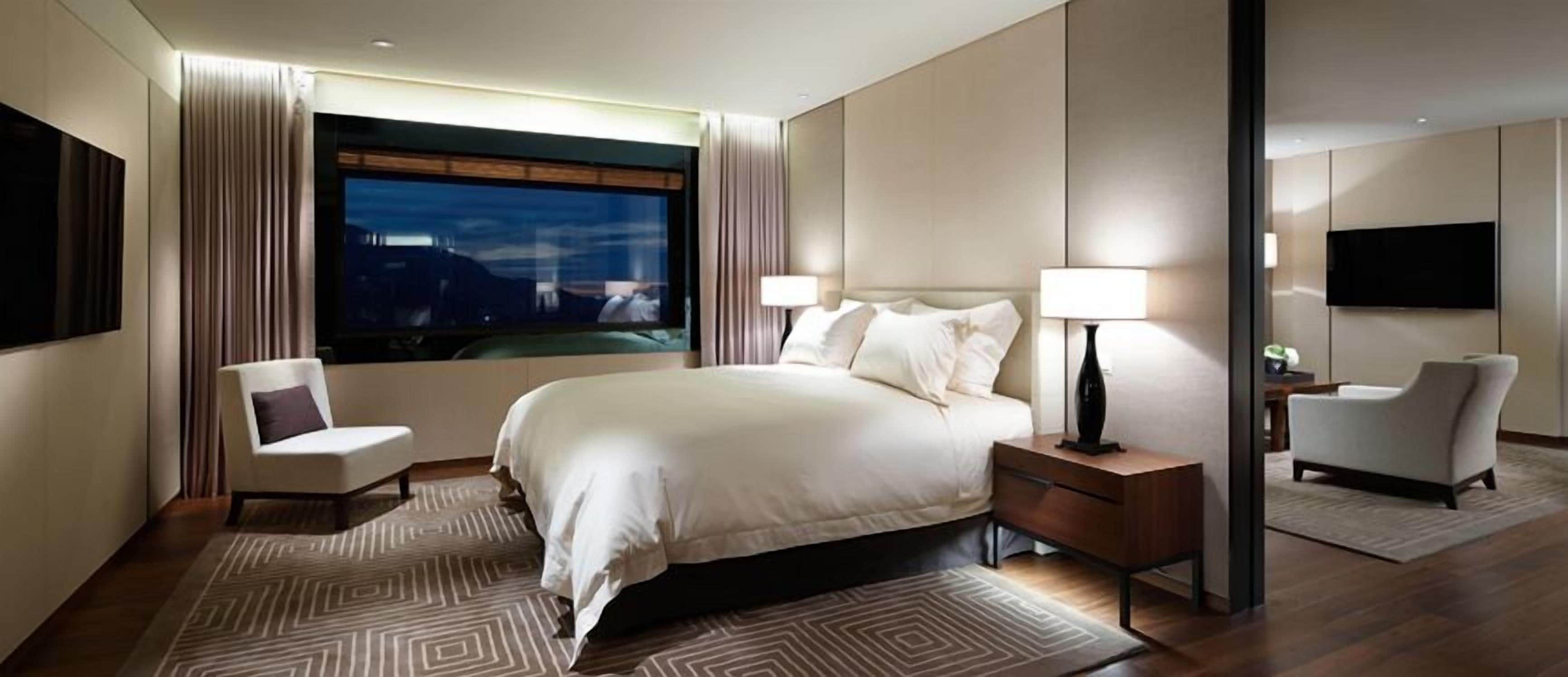 A modern hotel room with a large bed, two bedside tables, a TV, and a view of the city at night.