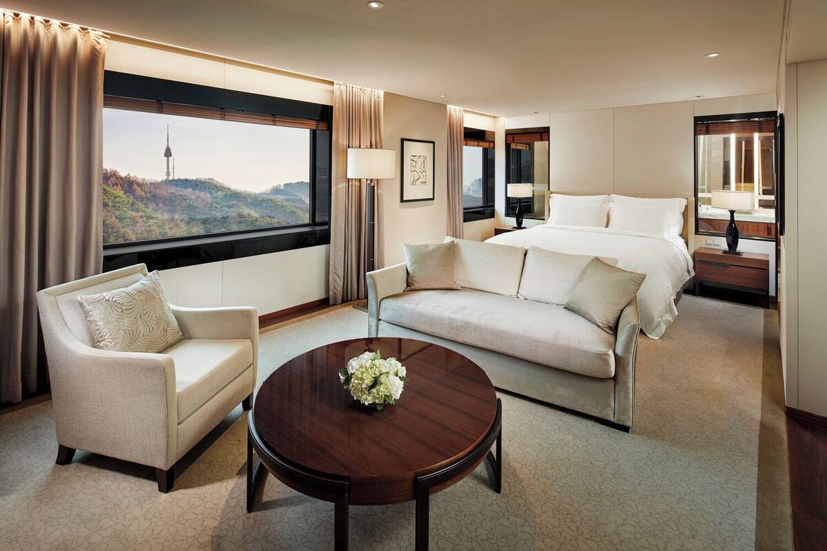 Quarto de hotel com uma cama grande, dois sofás, uma mesa de centro e vista para uma torre e montanhas.