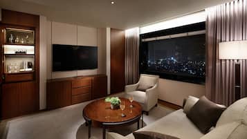 A modern living room with a flat-screen TV, a round wooden coffee table with a plant, a sofa, and a chair. There is a large window with a city view.