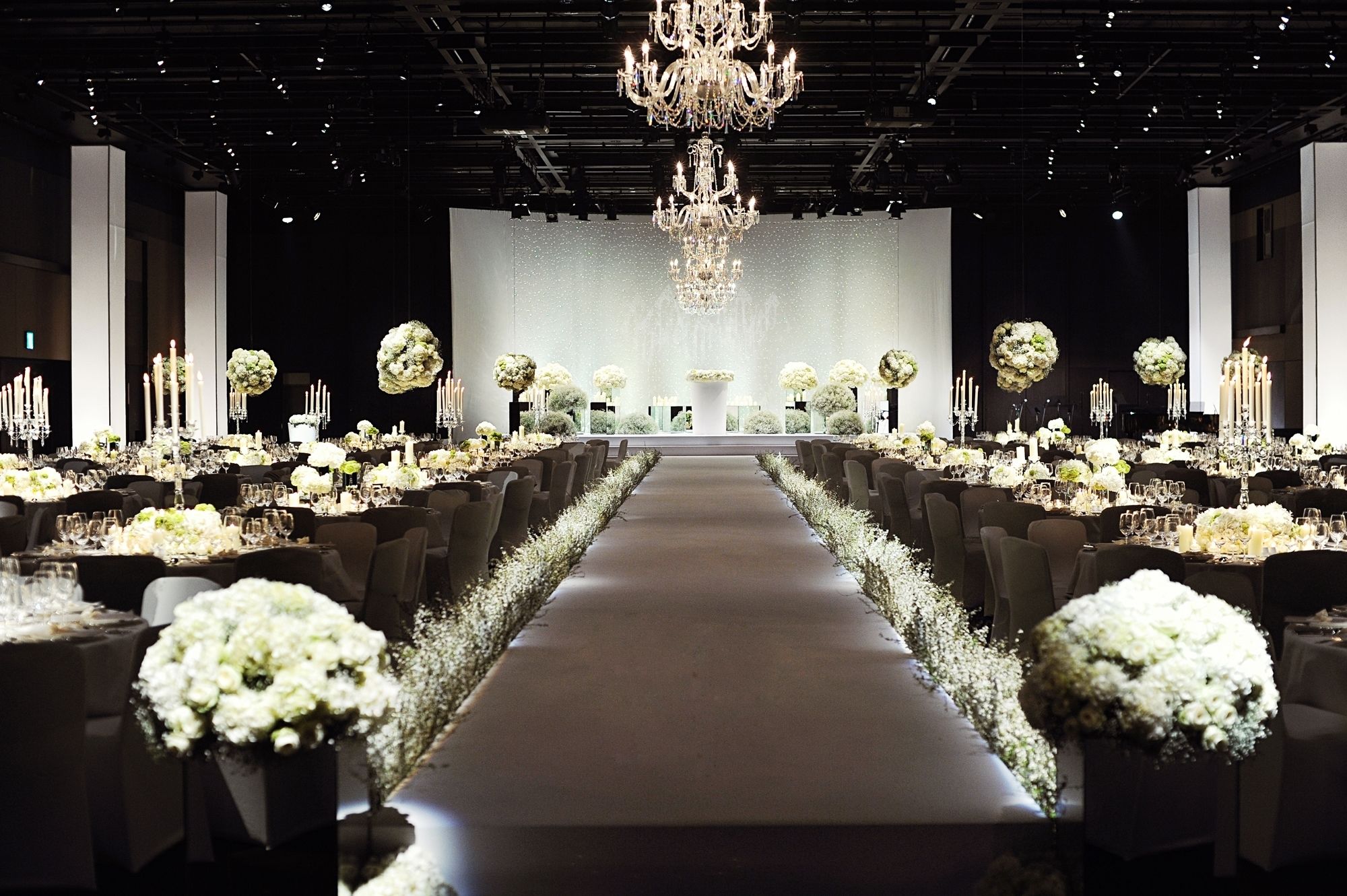 A long, narrow hall with rows of tables set for a formal event, each table adorned with white flowers and candles.