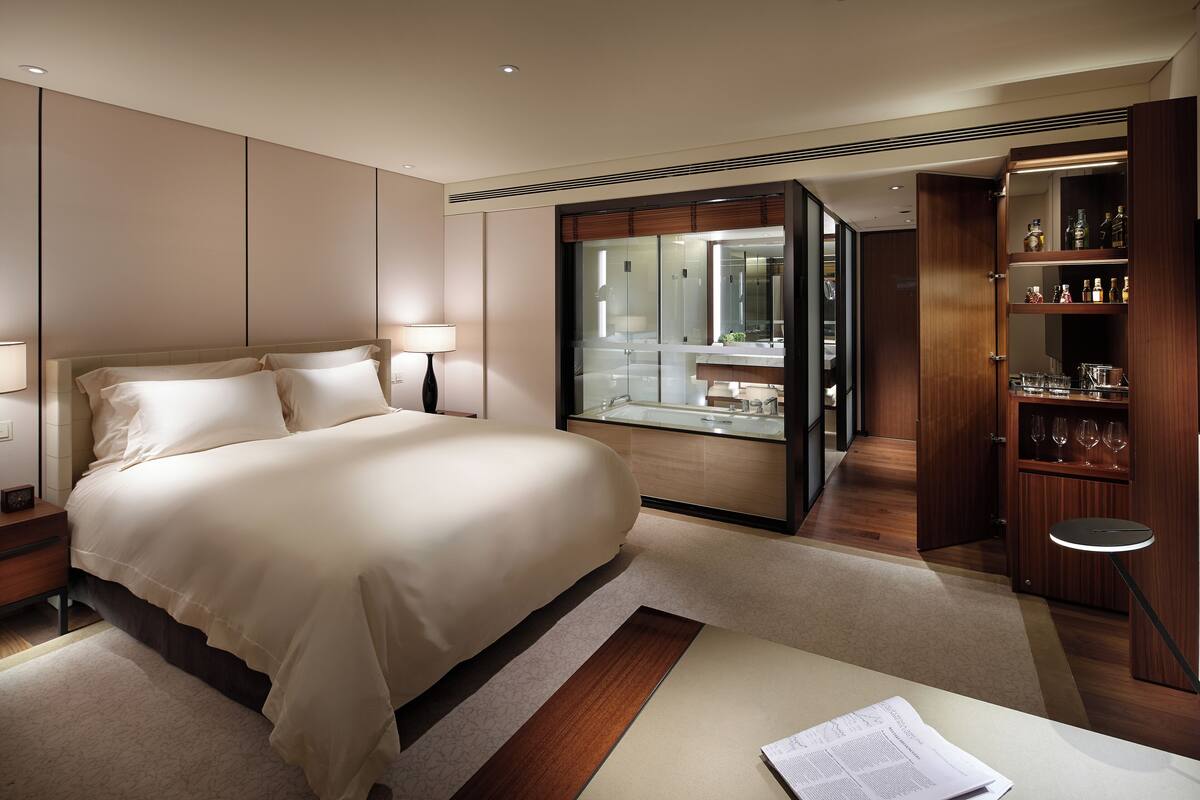 A modern hotel room with a large bed, a glass-enclosed bathroom, and a wooden cabinet with glassware.