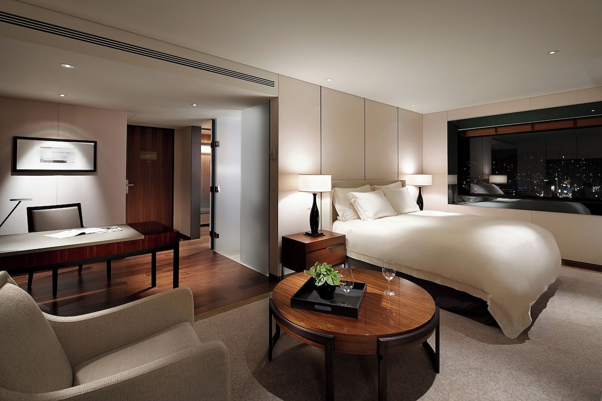 Une chambre d’hôtel moderne, équipée d’un lit, de lampes de chevet, d’un bureau et offrant une vue sur la ville la nuit.