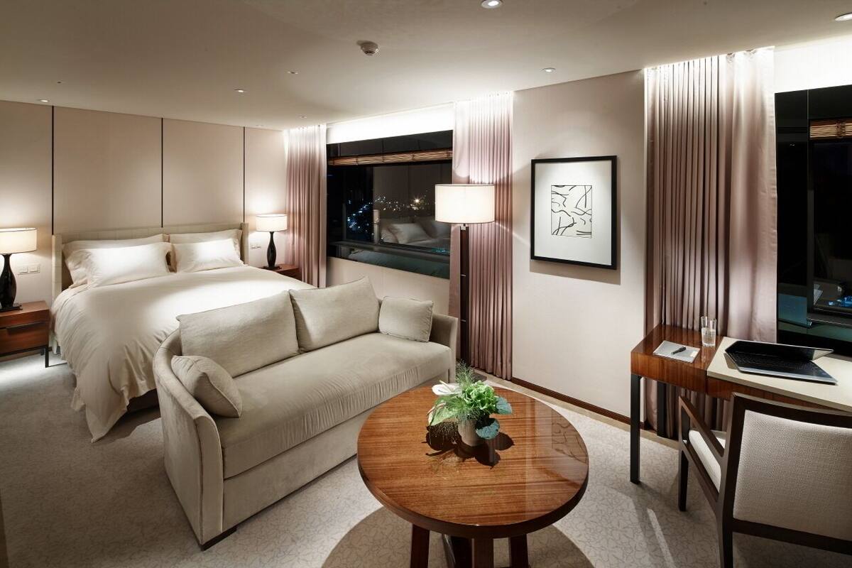 Quarto de hotel com cama, sofá, mesa redonda de madeira com uma planta, escrivaninha com um laptop e janela com cortinas.